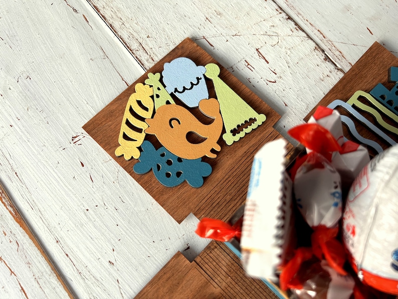 May include: A wooden box with a colorful birthday theme. The box is decorated with a bird, a cupcake, a party hat, and a striped candy. The box is filled with wrapped candies.