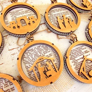 May include: Wooden Christmas ornaments with Bible verses and Nativity scene cutouts. Each ornament features a circular frame with a star-shaped top and a decorative border. The ornaments are made of light brown wood and are strung with twine.