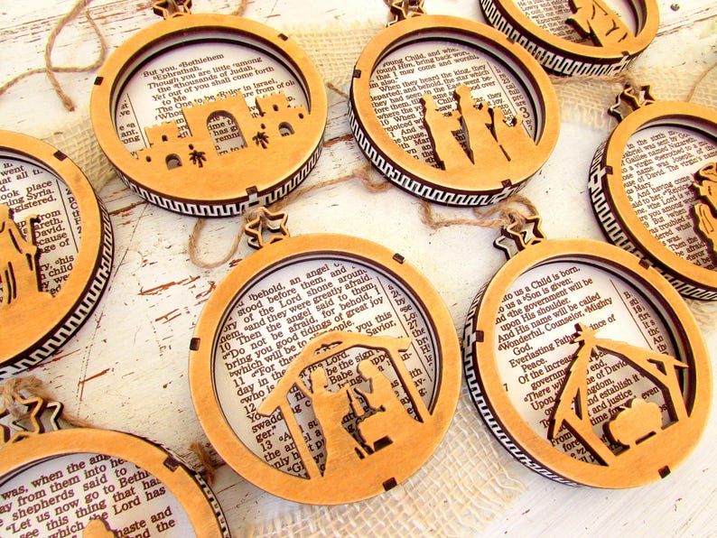 May include: Wooden Christmas ornaments with Bible verses and Nativity scene cutouts. Each ornament features a circular frame with a star-shaped top and a decorative border. The ornaments are made of light brown wood and are strung with twine.