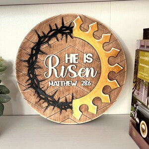May include: A round wooden sign with a brown background and a chevron pattern. The sign features a black crown of thorns and a gold crown. The text "He is Risen" and "Matthew 28:6" is written in white on the sign.