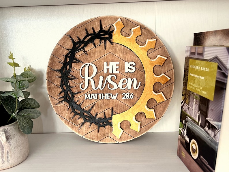 May include: A round wooden sign with a brown background and a chevron pattern. The sign features a black crown of thorns and a gold crown. The text "He is Risen" and "Matthew 28:6" is written in white on the sign.