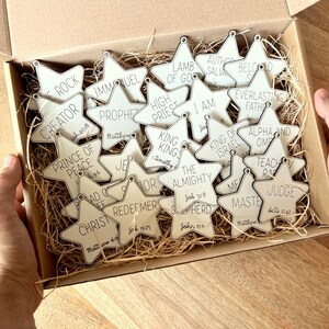 May include: A box filled with wooden star ornaments, each with a different biblical title and scripture reference. The titles include "Creator", "Prince of Peace", "Immanuel", "Prophet", "High Priest", "King of Kings", "The Almighty", "Shepherd", "Author and Savior", "Beloved", "Everlasting Father", "King of Israel", "Alpha and Omega", "Teacher", "Master", and "Judge".