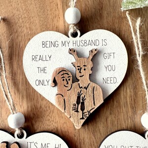 May include: White wooden heart-shaped ornament with a brown wood cutout of a couple holding champagne glasses. The ornament has the text "Being my husband is really the only gift you need."