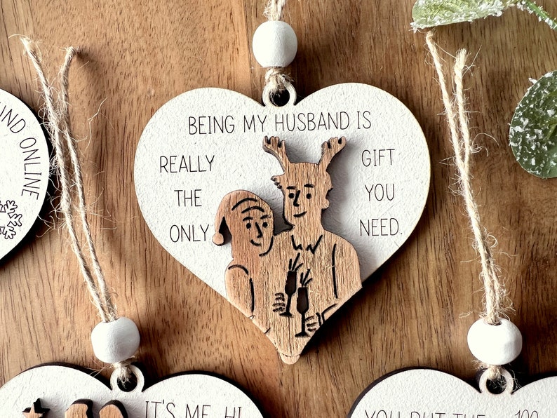 May include: White wooden heart-shaped ornament with a brown wood cutout of a couple holding champagne glasses. The ornament has the text "Being my husband is really the only gift you need."