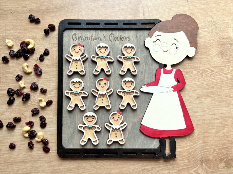 K&ouml;nnte beinhalten: Ein Holz-Lebkuchen-Backset mit einer Frau, die einen Teller h&auml;lt. Das Set enth&auml;lt acht Lebkuchen mit Namen wie "Oliver", "Sarah" und "Jack". Das Set ist mit "Grandma's Cookies" beschriftet.