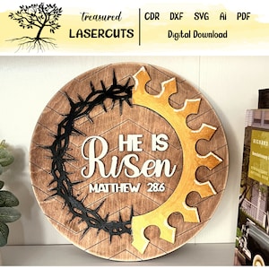 May include: A round wooden sign with a brown background and a chevron pattern. The sign features a black crown of thorns, a gold gear, and white text that reads "He is Risen Matthew 28:6".