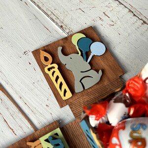 May include: A wooden box with a cutout of an elephant holding balloons and a candle. The box is filled with chocolate candies, including Kinder Country and Kinder Surprise.