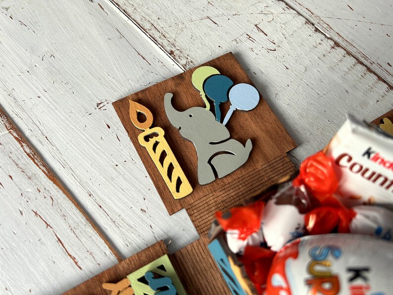 May include: A wooden box with a cutout of an elephant holding balloons and a candle. The box is filled with chocolate candies, including Kinder Country and Kinder Surprise.