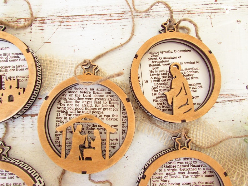 May include: Wooden Christmas ornaments with a circular design, featuring cut-out silhouettes of religious figures and scenes. Each ornament has a star-shaped top and is strung with twine. The background includes text from a book.