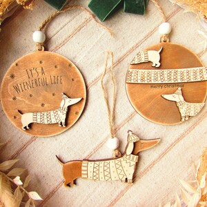 May include: Wooden Christmas ornaments featuring dachshund dogs wearing sweaters. One ornament reads "It's a Wienerful Life." Another says "merry christmas." Each ornament has a twine loop for hanging and a white bead.