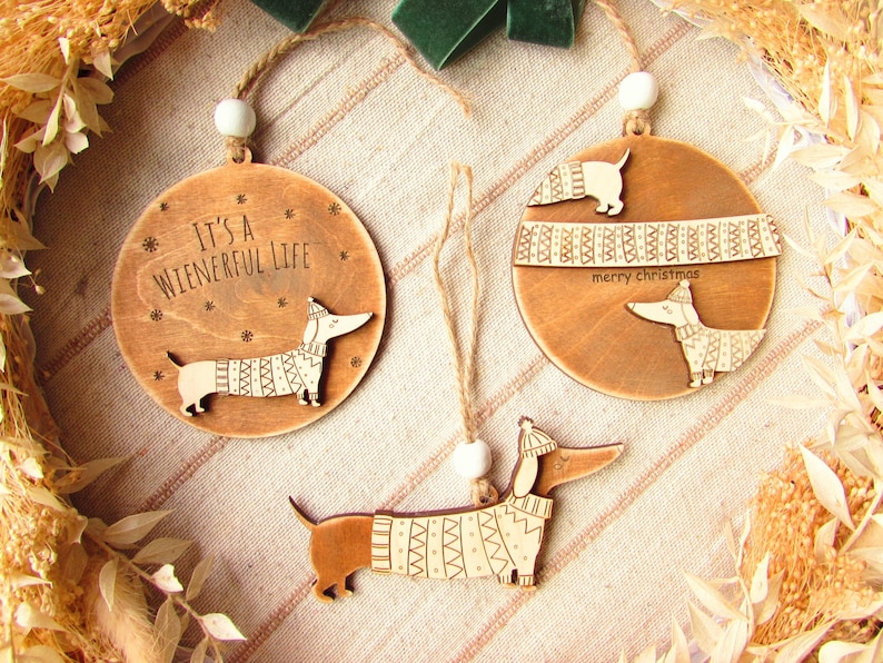 May include: Wooden Christmas ornaments featuring dachshund dogs wearing sweaters. One ornament reads "It's a Wienerful Life." Another says "merry christmas." Each ornament has a twine loop for hanging and a white bead.