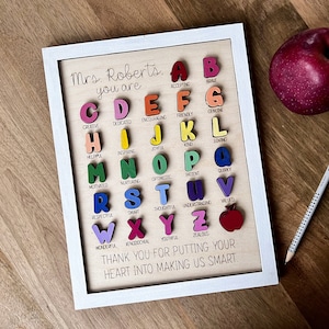 May include: A framed wooden sign with a white frame and a light brown background. The sign features a colourful alphabet with a word describing each letter. The sign reads "Mrs. Roberts. You are..." and ends with "Thank you for putting your heart into making us smart."