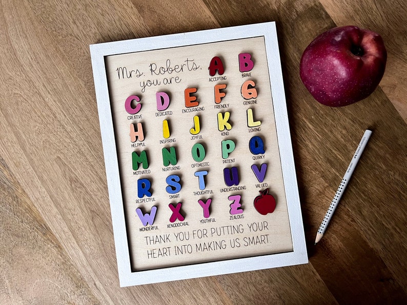 May include: A framed wooden sign with a white frame and a light brown background. The sign features a colourful alphabet with a word describing each letter. The sign reads "Mrs. Roberts. You are..." and ends with "Thank you for putting your heart into making us smart."
