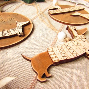 May include: Wooden dachshund ornaments with engraved details. One ornament is round with the text "It's a wienerful life" and a dachshund wearing a sweater. Other ornaments feature dachshunds with sweater designs.