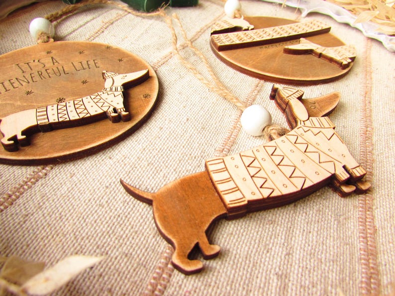 May include: Wooden dachshund ornaments with engraved details. One ornament is round with the text "It's a wienerful life" and a dachshund wearing a sweater. Other ornaments feature dachshunds with sweater designs.