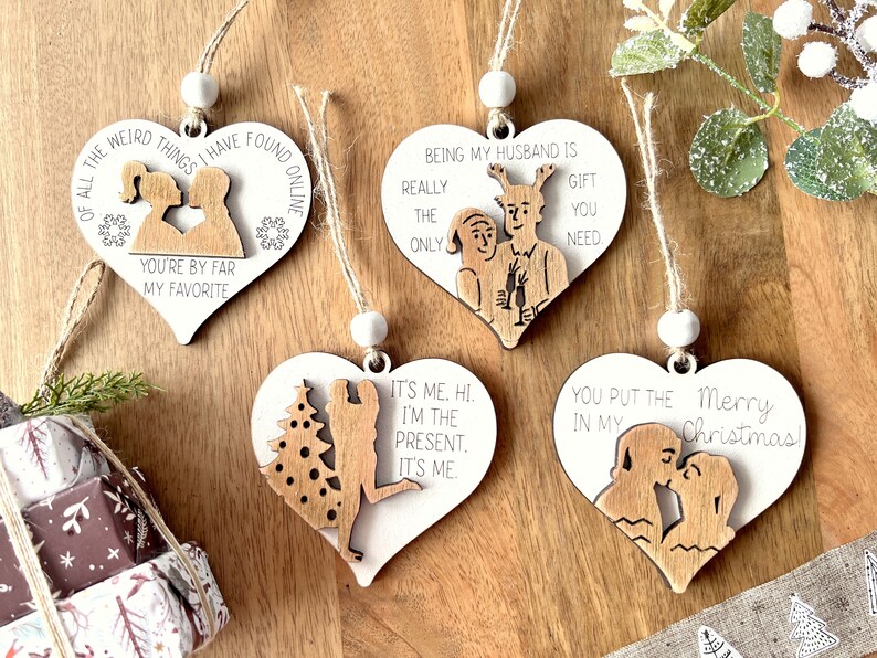 May include: Four wooden heart-shaped ornaments with different sayings and illustrations. The ornaments are white with brown wood accents. The sayings include "Of all the weird things I have found online, you're by far my favorite", "Being my husband is really the only gift you need", "It's me. Hi. I'm the present. It's me." and "You put the Merry Christmas in my".
