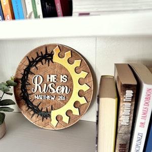 May include: A round wooden sign with a brown background and a gold crown design. The sign says "He is Risen" with the scripture reference "Matthew 28:6" underneath. The crown is surrounded by a black thorn wreath.
