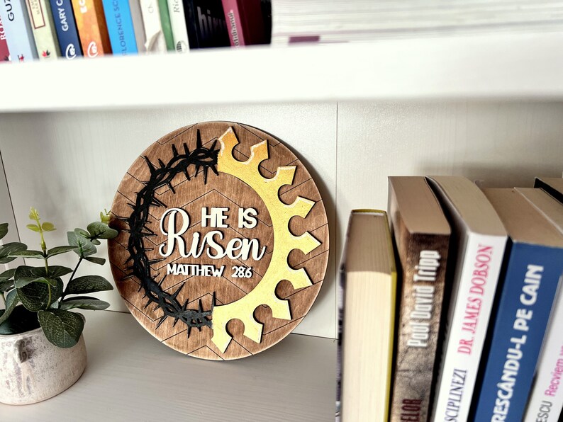 May include: A round wooden sign with a brown background and a gold crown design. The sign says "He is Risen" with the scripture reference "Matthew 28:6" underneath. The crown is surrounded by a black thorn wreath.