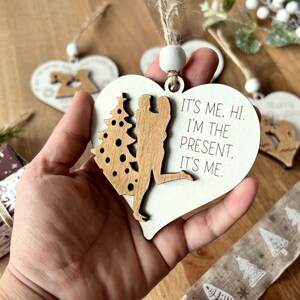 May include: White wooden heart-shaped ornament with a cutout of a couple standing under a Christmas tree. The ornament has the text "It's me. Hi. I'm the present. It's me."