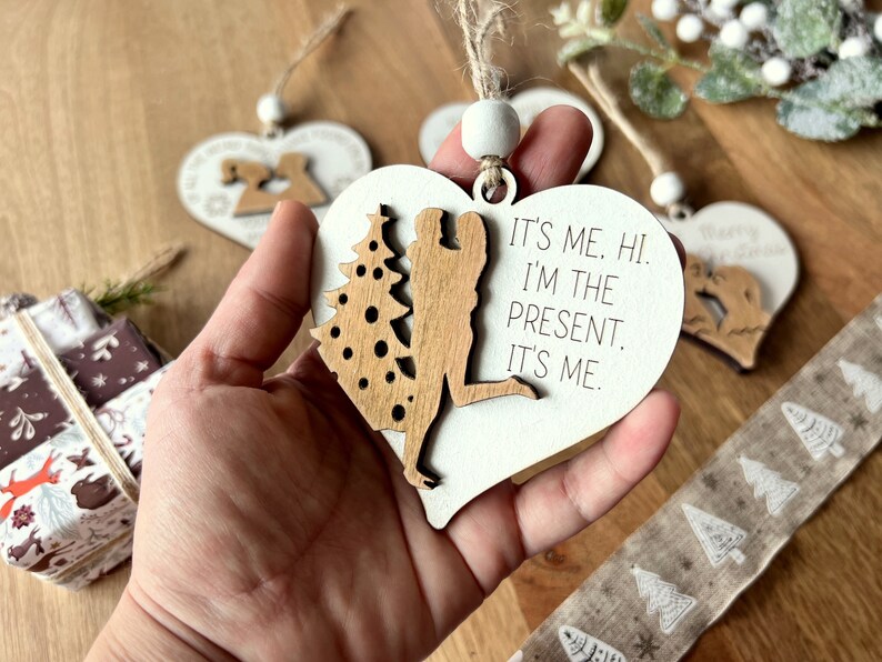 May include: White wooden heart-shaped ornament with a cutout of a couple standing under a Christmas tree. The ornament has the text "It's me. Hi. I'm the present. It's me."