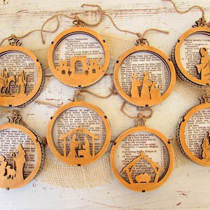 May include: A collection of eight wooden Christmas ornaments. Each ornament features a circular frame with a cutout design depicting a Nativity scene. The ornaments are strung with twine and have a star-shaped top. The background of each ornament is a page from a book.