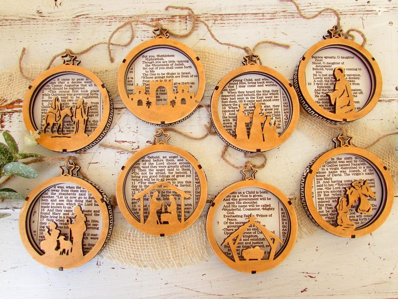 May include: A collection of eight wooden Christmas ornaments. Each ornament features a circular frame with a cutout design depicting a Nativity scene. The ornaments are strung with twine and have a star-shaped top. The background of each ornament is a page from a book.