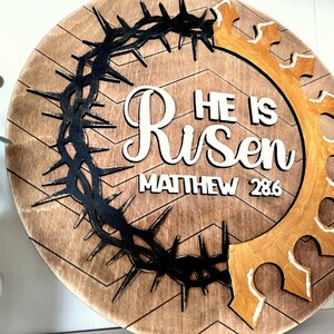 May include: A round wooden sign with a brown chevron pattern. The sign features a black crown of thorns, a gold crown, and white lettering that reads "He is Risen Matthew 28:6".