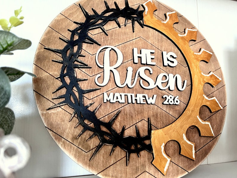 May include: A round wooden sign with a brown chevron pattern. The sign features a black crown of thorns, a gold crown, and white lettering that reads "He is Risen Matthew 28:6".