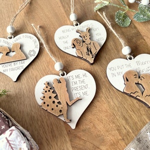May include: Set of four wooden heart-shaped Christmas ornaments with various sayings and illustrations. The ornaments feature sayings like "You're by far my favorite", "Being my husband is the only gift you need", "It's me. Hi. I'm the present. It's me", and "You put the Merry Christmas in my".