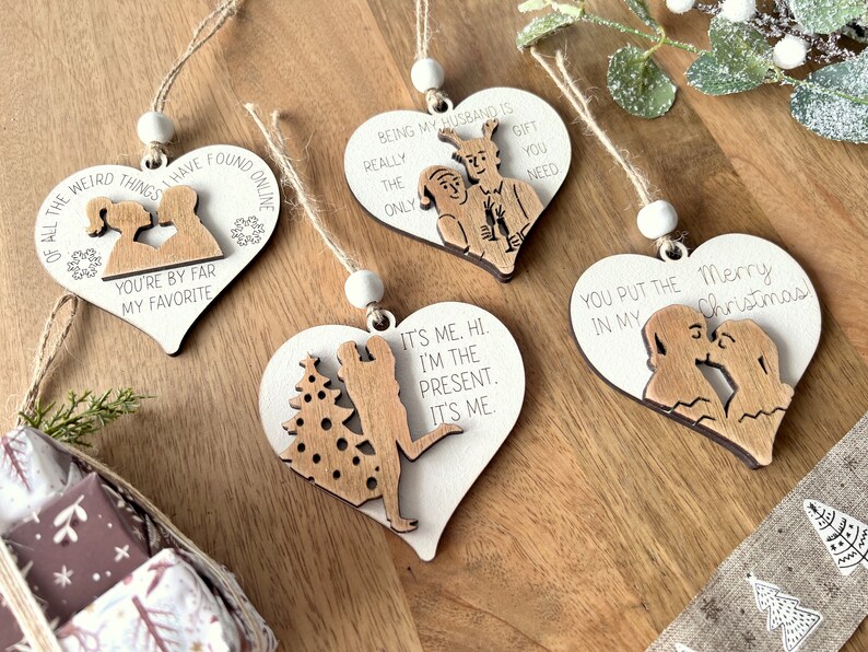 May include: Set of four wooden heart-shaped Christmas ornaments with various sayings and illustrations. The ornaments feature sayings like "You're by far my favorite", "Being my husband is the only gift you need", "It's me. Hi. I'm the present. It's me", and "You put the Merry Christmas in my".