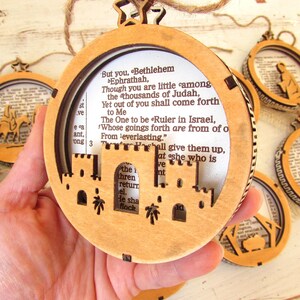 May include: Wooden Christmas ornaments with a circular design. The ornaments feature a laser-cut Bethlehem scene and text from the Bible. Each ornament is suspended by a twine loop and a star-shaped top.