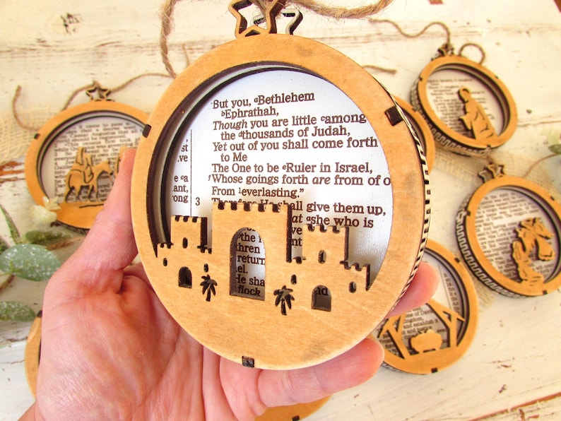 May include: Wooden Christmas ornaments with a circular design. The ornaments feature a laser-cut Bethlehem scene and text from the Bible. Each ornament is suspended by a twine loop and a star-shaped top.