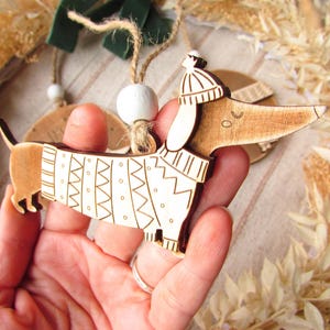 May include: Wooden dachshund ornament with a white sweater featuring a geometric pattern and a matching hat. The dog has a string for hanging. A festive holiday decoration.