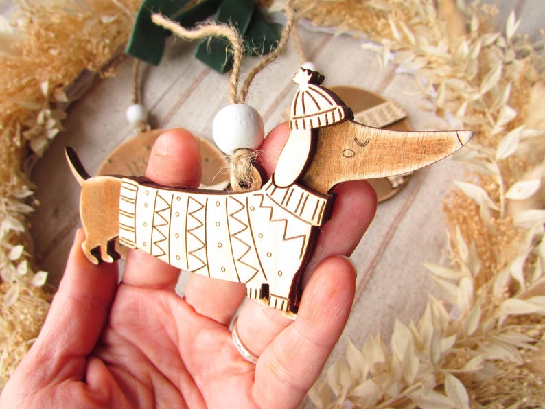 May include: Wooden dachshund ornament with a white sweater featuring a geometric pattern and a matching hat. The dog has a string for hanging. A festive holiday decoration.