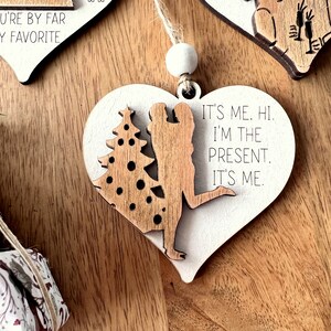 May include: White heart-shaped wooden ornament with a couple silhouette and a Christmas tree. The ornament has the text "It's me. Hi. I'm the present. It's me."