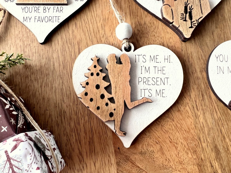 May include: White heart-shaped wooden ornament with a couple silhouette and a Christmas tree. The ornament has the text "It's me. Hi. I'm the present. It's me."