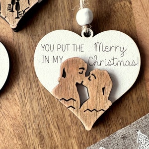 May include: A white wooden heart-shaped ornament with a couple kissing inside. The ornament has the text "You put the Merry Christmas!" on it.