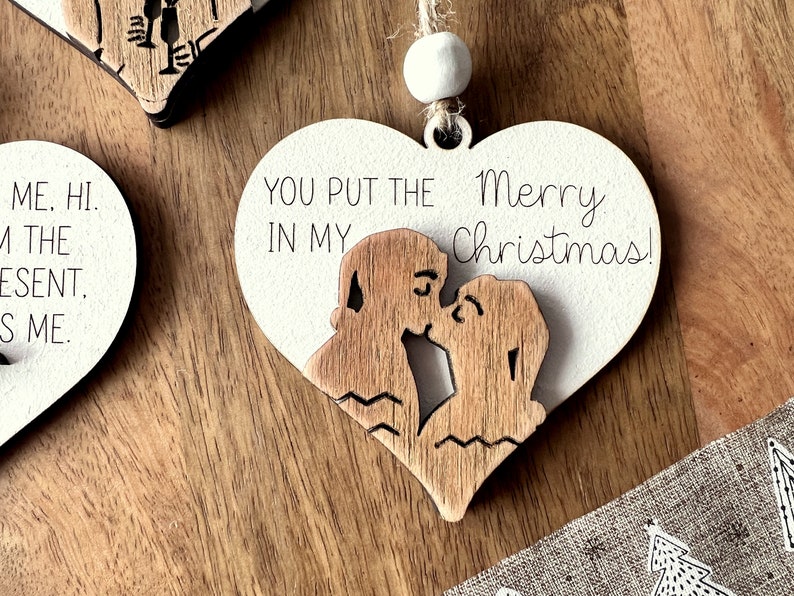 May include: A white wooden heart-shaped ornament with a couple kissing inside. The ornament has the text "You put the Merry Christmas!" on it.