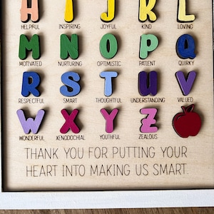 May include: A wooden alphabet chart with colourful letters and words describing positive character traits. The words include helpful, inspiring, joyful, kind, loving, motivated, nurturing, optimistic, patient, quirky, respectful, smart, thoughtful, understanding, valued, wonderful, xenodochial, youthful, and zealous. The chart also includes a red apple and the text "THANK YOU FOR PUTTING YOUR HEART INTO MAKING US SMART."