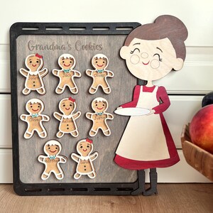 K&ouml;nnte beinhalten: Ein Holz-Lebkuchen-Adventskalender mit einer Lebkuchenfrau, die einen Teller h&auml;lt. Der Kalender zeigt acht Lebkuchenm&auml;nner mit Namen wie "Janet", "Jack", "Oliver", "Melissa", "Justin", "Oliver", "Matthew" und "Sarah". Der Kalender tr&auml;gt den Titel "Grandma's Cookies".