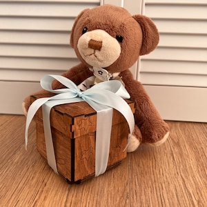 May include: A brown teddy bear holding a wooden gift box with a blue ribbon. The box has a decorative design with stars and other shapes.