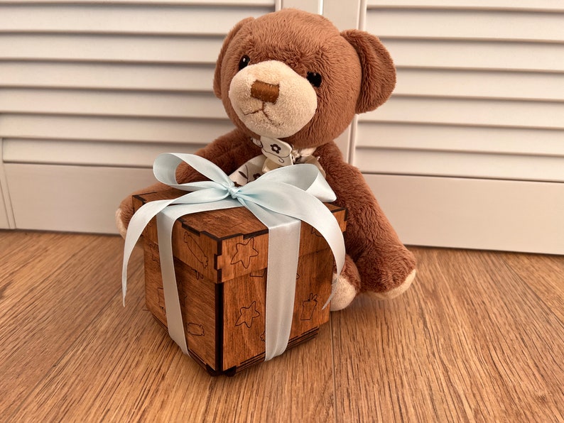May include: A brown teddy bear holding a wooden gift box with a blue ribbon. The box has a decorative design with stars and other shapes.