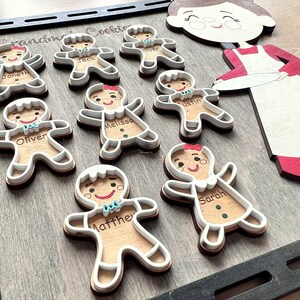 K&ouml;nnte beinhalten: Ein Holz-Lebkuchen-Kekse-Dekorationsset mit acht Lebkuchenmann-Formen, jede mit einem anderen Namen. Das Set enth&auml;lt au&szlig;erdem eine gr&ouml;&szlig;ere Lebkuchenmann-Form mit einem rot-wei&szlig;en Kleid und einem Teller in den H&auml;nden. Der Text "Grandma's Cookies" ist auf dem Holzbrett geschrieben.