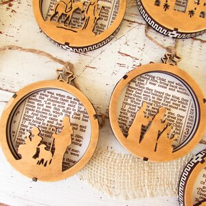 May include: Wooden Christmas ornaments with a religious theme. The ornaments feature a laser-cut design of the nativity scene, with a star-shaped top and a twine hanger. The background is a page from a book.
