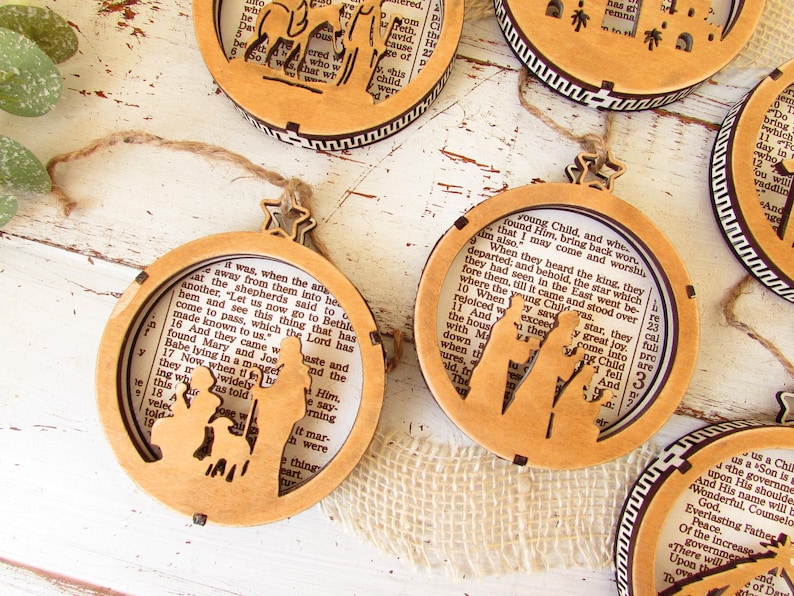 May include: Wooden Christmas ornaments with a religious theme. The ornaments feature a laser-cut design of the nativity scene, with a star-shaped top and a twine hanger. The background is a page from a book.