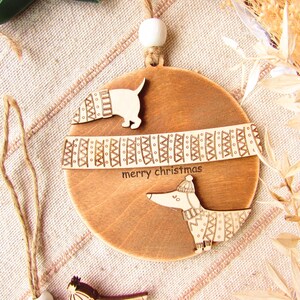 May include: Wooden Christmas ornament featuring a dachshund dog design. The ornament is round with a light brown finish and includes the words "merry christmas". The dog is wearing a hat and scarf.