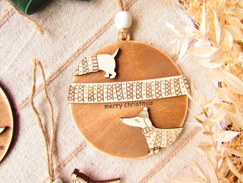 May include: Wooden Christmas ornament featuring a dachshund dog design. The ornament is round with a light brown finish and includes the words "merry christmas". The dog is wearing a hat and scarf.
