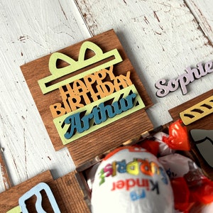 May include: A wooden box with a colorful "Happy Birthday Arthur" sign, a yellow candle, a gray elephant, and a pink "Sophie" name tag. The box is open and contains a red and white candy.