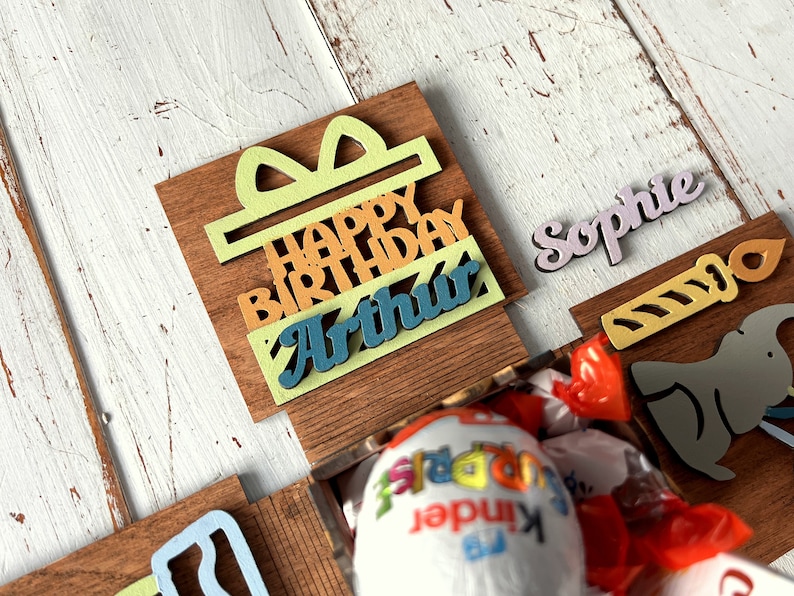 May include: A wooden box with a colorful "Happy Birthday Arthur" sign, a yellow candle, a gray elephant, and a pink "Sophie" name tag. The box is open and contains a red and white candy.
