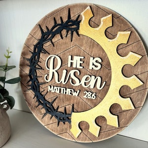 May include: A round wooden sign with a brown wood background and a chevron pattern. The sign features a black crown of thorns, a gold crown, and the words "He is Risen" with the scripture reference "Matthew 28:6".